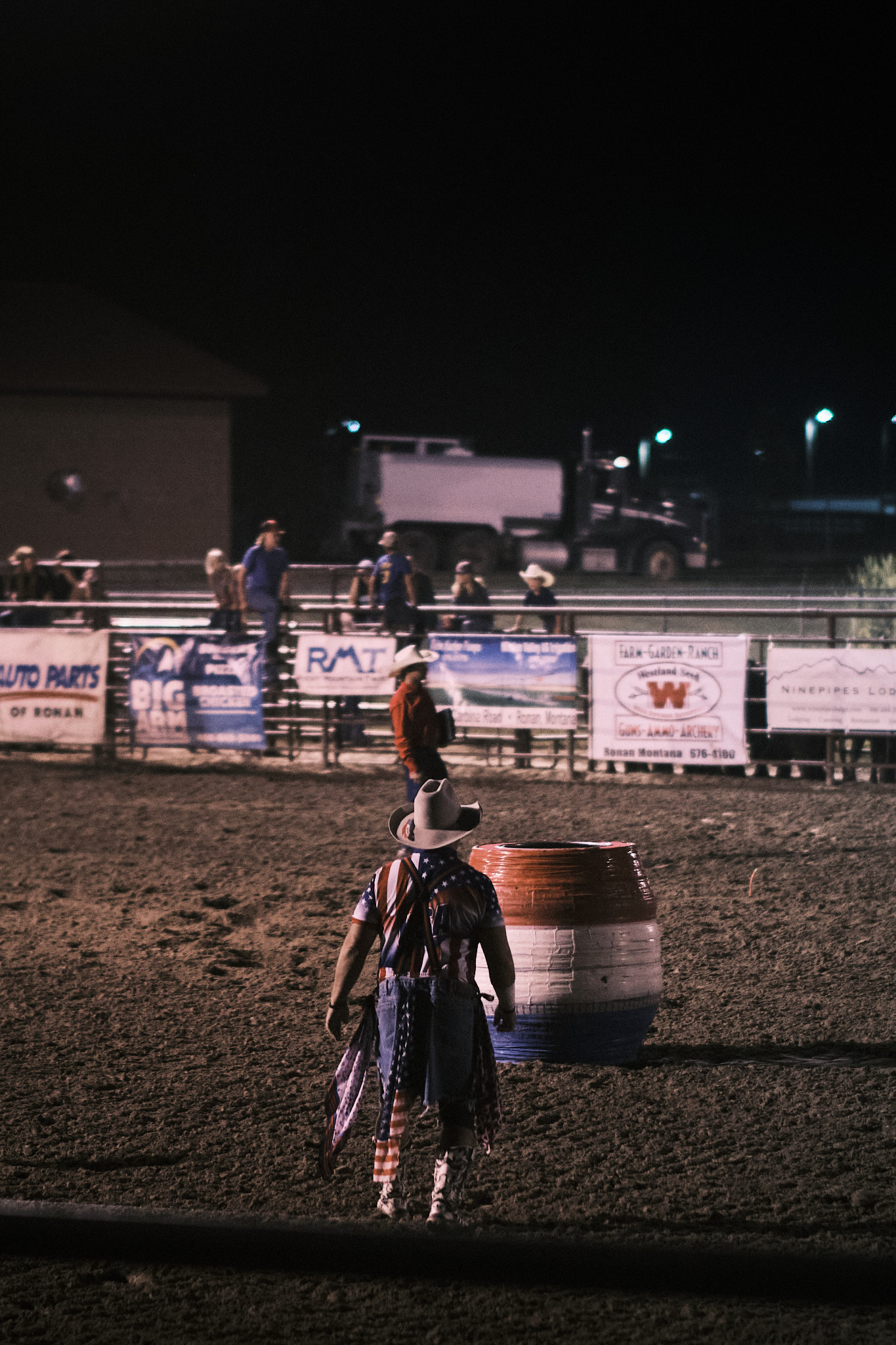 A cowboy in a red, white, and blue outfit standing in the arena by a red, white, and blue barrel