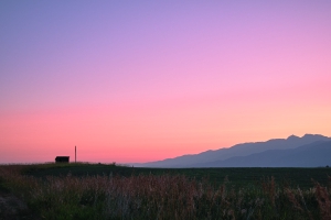 An early morning sunrise, a small structure on a green hill, the purple mountains off to the right, the sky a spectrum of pinks, reds, yellows, and purples