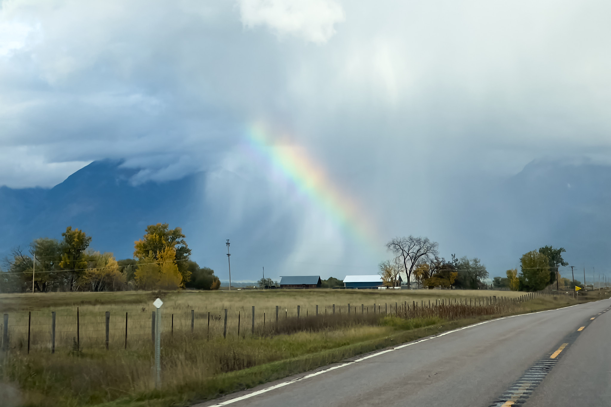 A rainbow beginning at a ranch house and rising toward a dying raincloud, the mountains peeking through behind them, the signs of early fall in the foreground