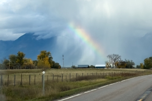 A rainbow beginning at a ranch house and rising toward a dying raincloud, the mountains peeking through behind them, the signs of early fall in the foreground